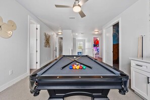 Een pooltafel in een gang met een plafondventilator, witte muren en een deur naar een andere kamer.