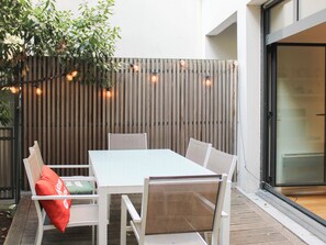 Un patio al aire libre con mesa y sillas blancas, una cerca de madera y luces colgantes.