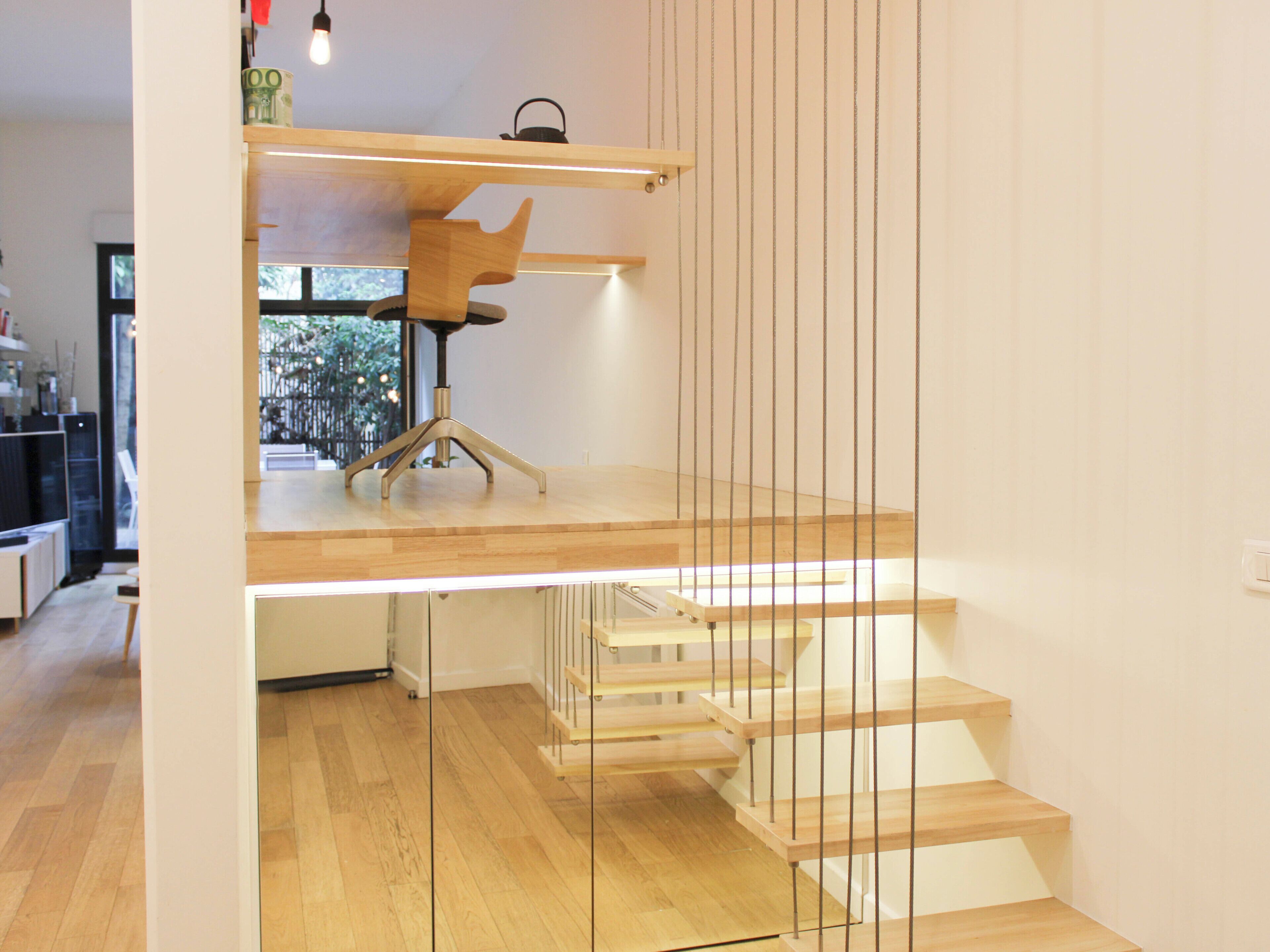 A modern interior with a wooden staircase, a desk with a chair, and a hanging light fixture.