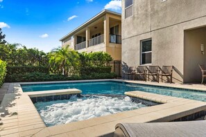 Piscina com hidromassagem, rodeada por espreguiçadeiras e um edifício com varandas.