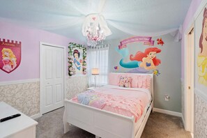A bedroom with a bed, a dresser, a chandelier, and wall decorations including a mermaid mural and a Disney princess shield.