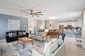A modern living room with a dark sofa, a light-colored sofa, and a wooden coffee table. There is a dining area with a table and chairs, and a kitchen with white cabinets and a stainless steel refrigerator. - Harry Potter Games Room/Full resort facilties includeded/Private Pool and HotTub (Lake Buena Vista)