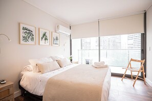 Une chambre à coucher avec un grand lit, une table de chevet, une commode, une fenêtre donnant sur des immeubles et un ventilateur de plafond.