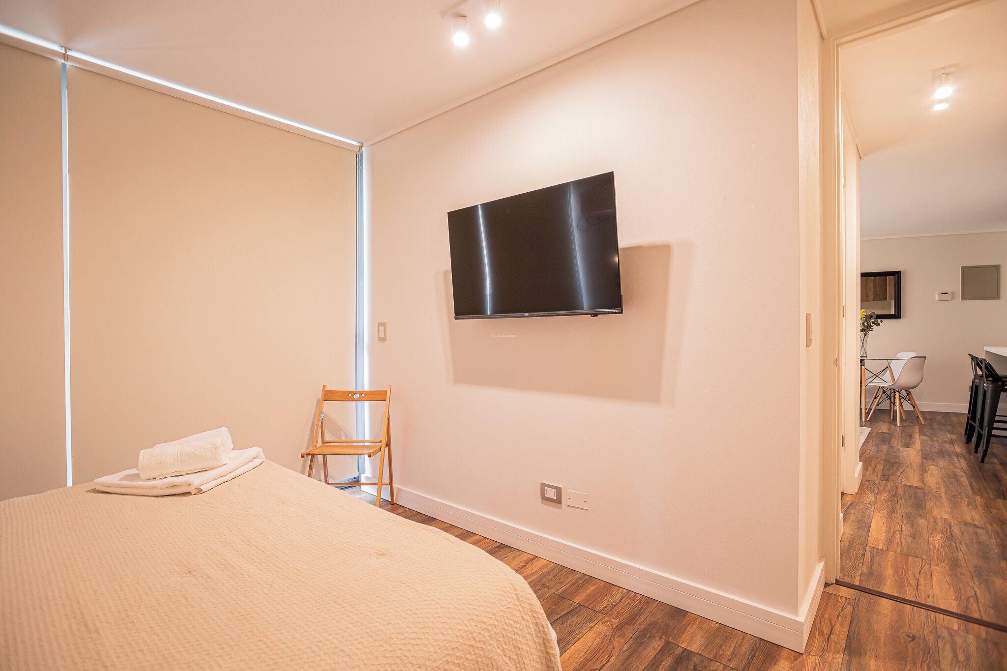 Un dormitorio con cama, una silla, un televisor de pantalla plana montado en la pared y piso de madera.