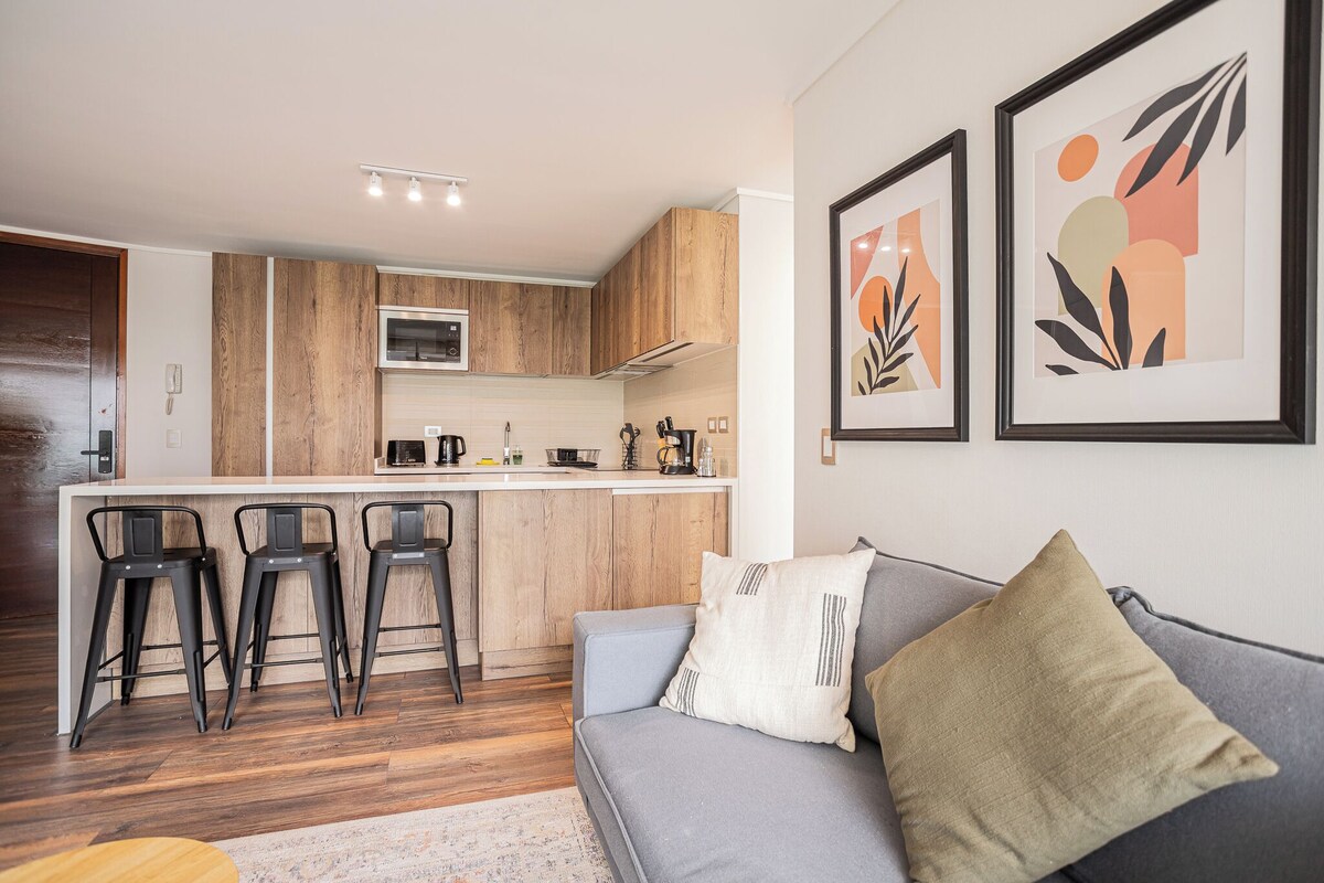 Eine moderne Küche mit Holdekor, einer Mikrowelle und einer Kaffeemaschine. Ein graues Sofa mit Kissen und ein kleiner Tisch mit Stühlen sind ebenfalls vorhanden.