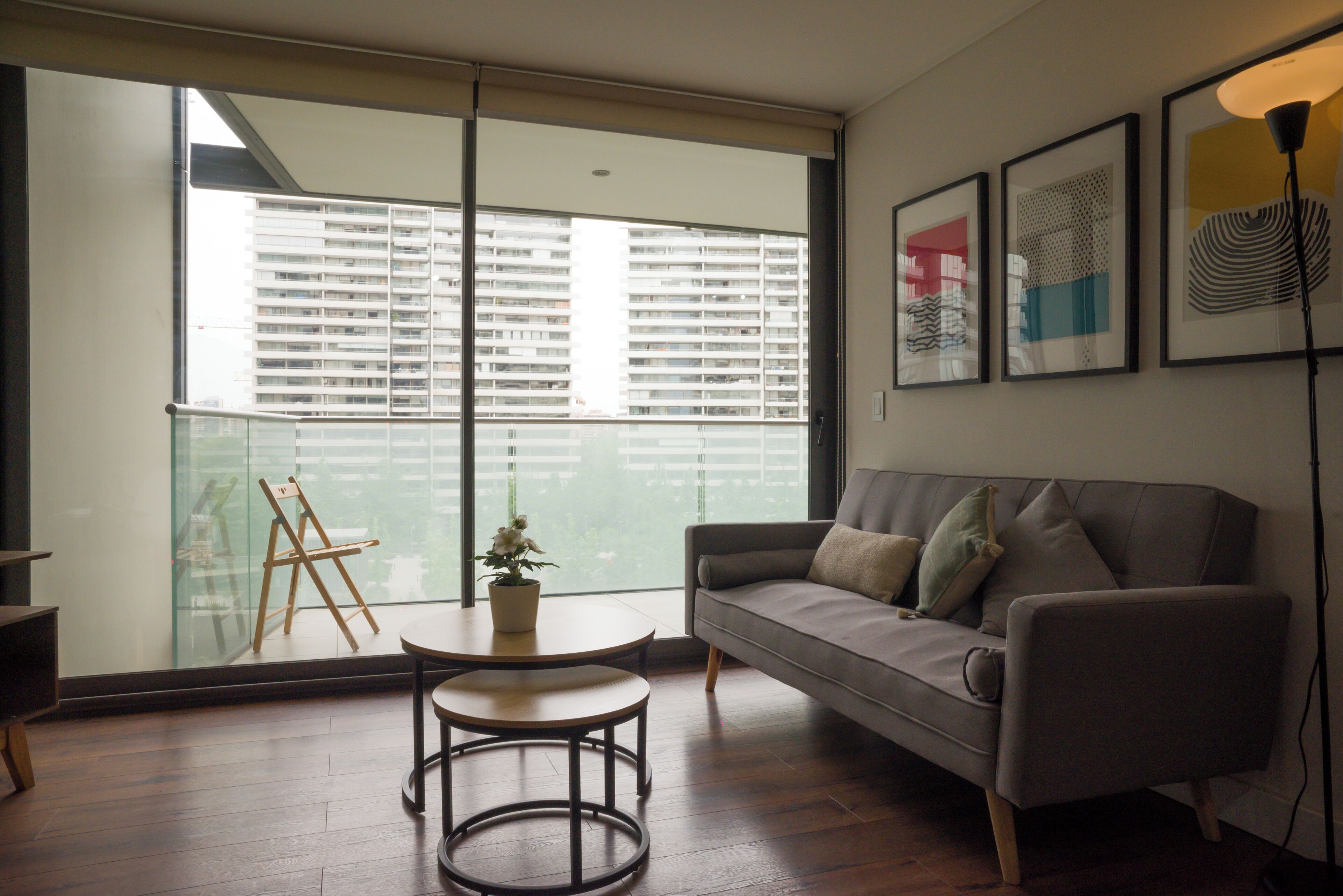 Una sala moderna con un sofá gris, una mesita redonda y una planta en maceta. Un ventanal grande ofrece vistas a un edificio de gran altura.