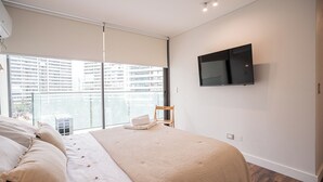 Ein modernes Schlafzimmer mit einem großen Bett, einem flachbildfernseher an der Wand und Blick auf ein Hochhaus durch ein großes Fenster.