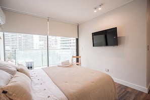 Ein modernes Schlafzimmer mit einem großen Bett, einem flachbildfernseher an der Wand und Blick auf ein Hochhaus durch ein großes Fenster.