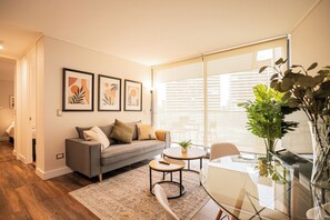 A modern living room with a sofa, coffee table, and chairs, a large window with blinds, and framed artwork on the wall. - Manquehue Norte Apartments by Andes STR (Las Condes)
