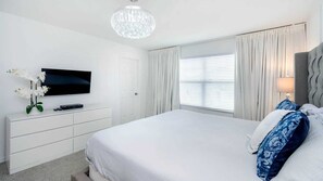 Un dormitorio de hotel con una cama grande, un televisor sobre una cómoda blanca, una araña y una ventana con cortinas.
