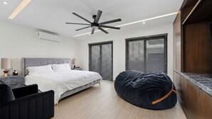 Ein modernes Schlafzimmer mit Bett, Deckenventilator, Sitzsack und großen Fenstern mit Jalousien.