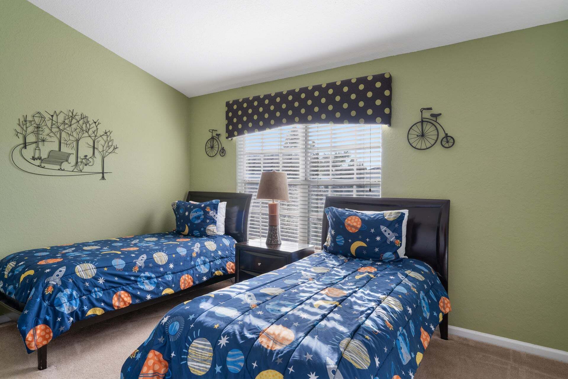 A bedroom with two beds, a window with blinds, a lamp, and wall art including a bicycle and a tree.