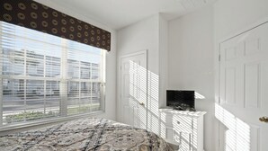 Een slaapkamer met een bed, een televisie op een witte dressoir, en een raam met zonwering.