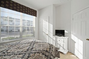A bedroom with a bed, a television on a white stand, and a window with blinds. - Firefly Cottage - Four Bedroom Apartment, Sleeps 8 (Kissimmee)