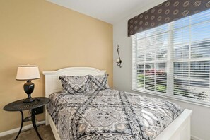 Een slaapkamer met een bed, een nachtkastje met lamp, een raam met jaloezieën en een wandlamp.
