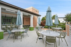 Ein Außenbereich mit einer Terrasse, runden Tischen und Stühlen, der von Sonnenschirmen beschattet wird, und ein Blick auf ein Gebäude im Hintergrund.