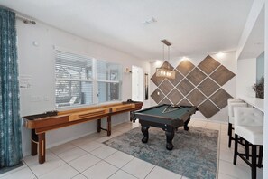 Una mesa de billar en una habitación con una ventana, una pared con diseño geométrico y una lámpara colgante.