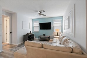 A living room with a beige sofa, a flat-screen TV mounted on the wall, a ceiling fan, and a window with blinds. - Galaxy Getaway - Nine Bedroom House, Sleeps 22 (Kissimmee)