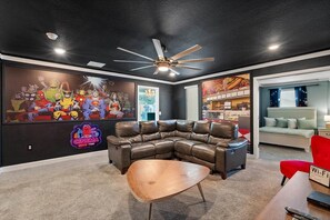 Ein Wohnzimmer mit einem ledernen Ecksofa, einem Holztisch, einem Deckenventilator, einer Wandmalerei und einem durch eine offene Tür einsehbarer Küchenbereich.
