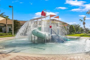 Ein Wasserspielplatz mit Rutsche und einem roten Eimer, umgeben von einer gepflasterten Fläche und Palmen.