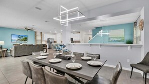 Un espace de repas moderne avec une longue table dressée pour six personnes, une cuisine en arrière-plan et un salon avec un canapé et un téléviseur.