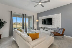 A modern living room with a white sofa, a dining table, a TV mounted on a textured wall, and a ceiling fan. - The Splash Shot -Championsgate Resort, Amenities Included/Games Room/Free wifi (Kissimmee)