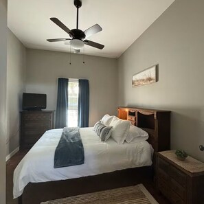 Une chambre à coucher comprenant un lit, un ventilateur de plafond, une commode, un téléviseur et une fenêtre avec des rideaux.