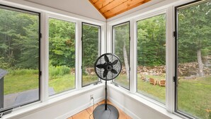 Een kamer met grote ramen, een ventilator en uitzicht op een bebost gebied.