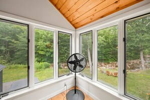 A room with large windows, a standing fan, and a view of a wooded area. - The Mad River Lodge: Riverside Escape (Waterville Valley)