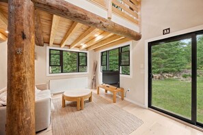 Ein Zimmer mit Holzboden, großen Fenstern, einem Fernseher auf einem Schrank und Blick auf eine bewaldete Landschaft.