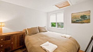 Een slaapkamer met een bed, een houten dressoir, een raam met jaloezieën en een schilderij aan de muur.