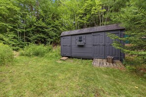 A small, dark-colored shed with a window and a door, surrounded by dense green foliage. - The Mad River Lodge: Riverside Escape (Waterville Valley)