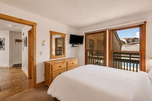Ein Schlafzimmer mit einem Bett, einer Holzkommode, einem Fernseher und einem großen Fenster mit Balkon.