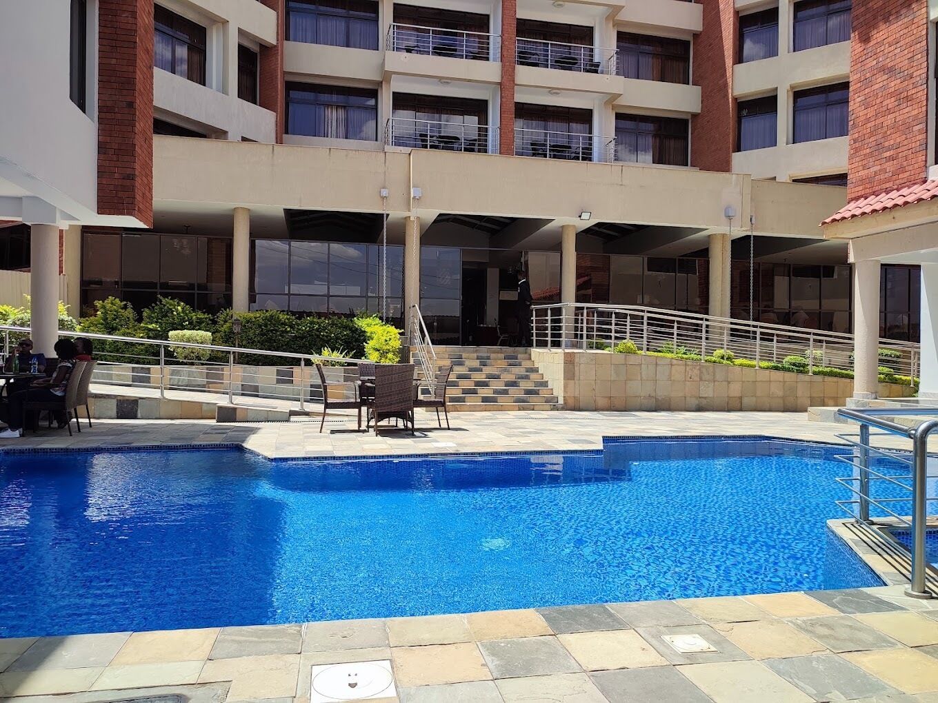 A hotel pool area with a clear blue pool, outdoor seating, and a staircase leading to the upper floors.