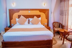Quarto de hotel com cama de madeira, duas luminárias de cabeceira, uma cadeira e uma mesa de cabeceira.