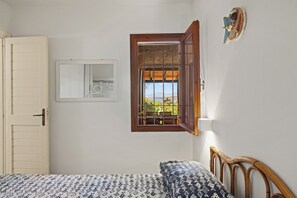Ein Zimmer mit einem Bett, einem Fenster mit geöffnetem Klappladen, einer Tür und einem an der Wand befestigten Spiegel.