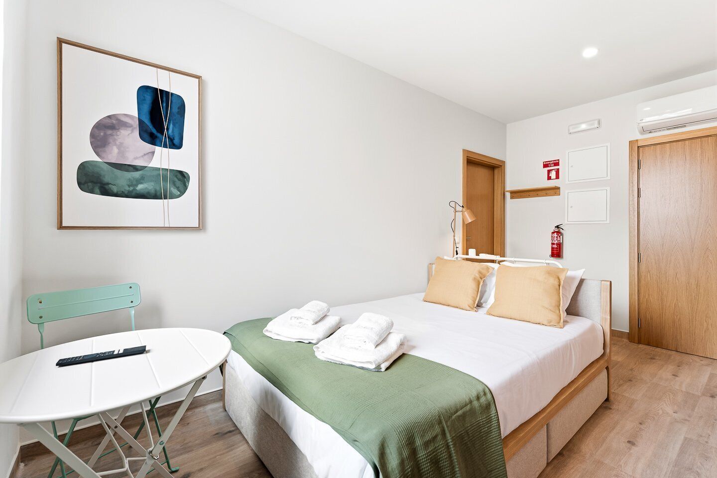 A hotel room with a bed, a small table, a chair, a framed abstract painting, and a fire extinguisher.
