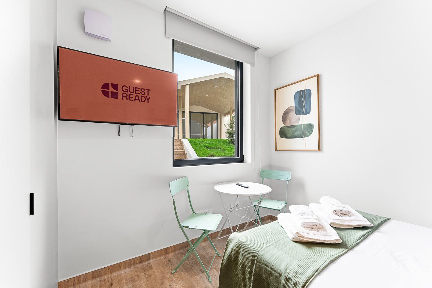 A hotel room with a bed, a desk with a chair, a small table with a chair, a TV mounted on the wall, and a window with a view of the outside.