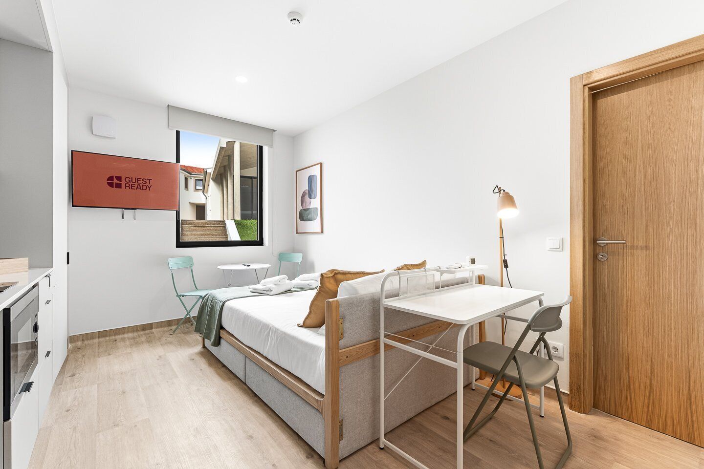 A compact hotel room with a bed, a small desk, a chair, a table, a TV, and a door.