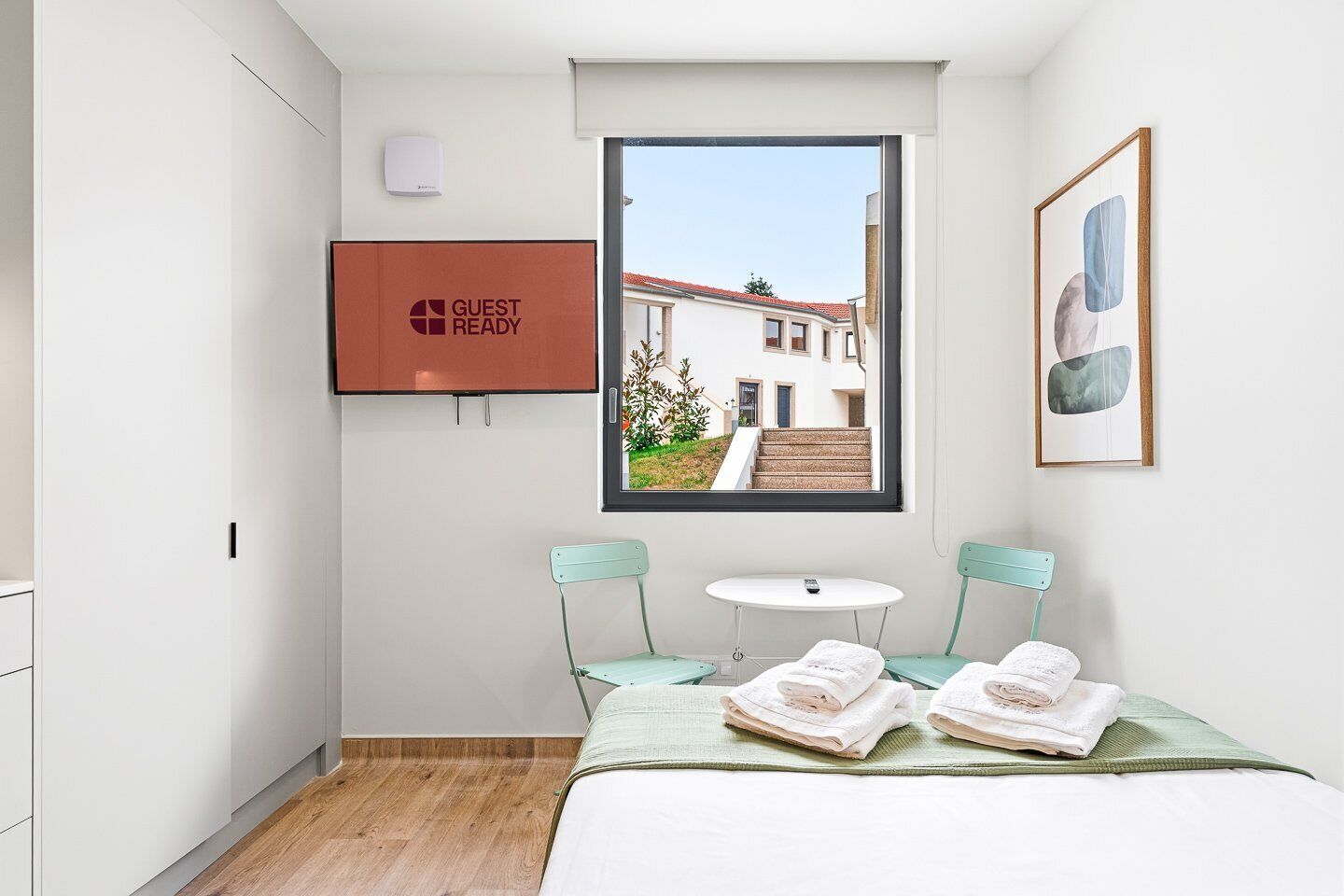 Une chambre d’hôtel avec un lit, deux chaises, une petite table, une fenêtre donnant sur des immeubles, un téléviseur et un tableau encadré au mur.