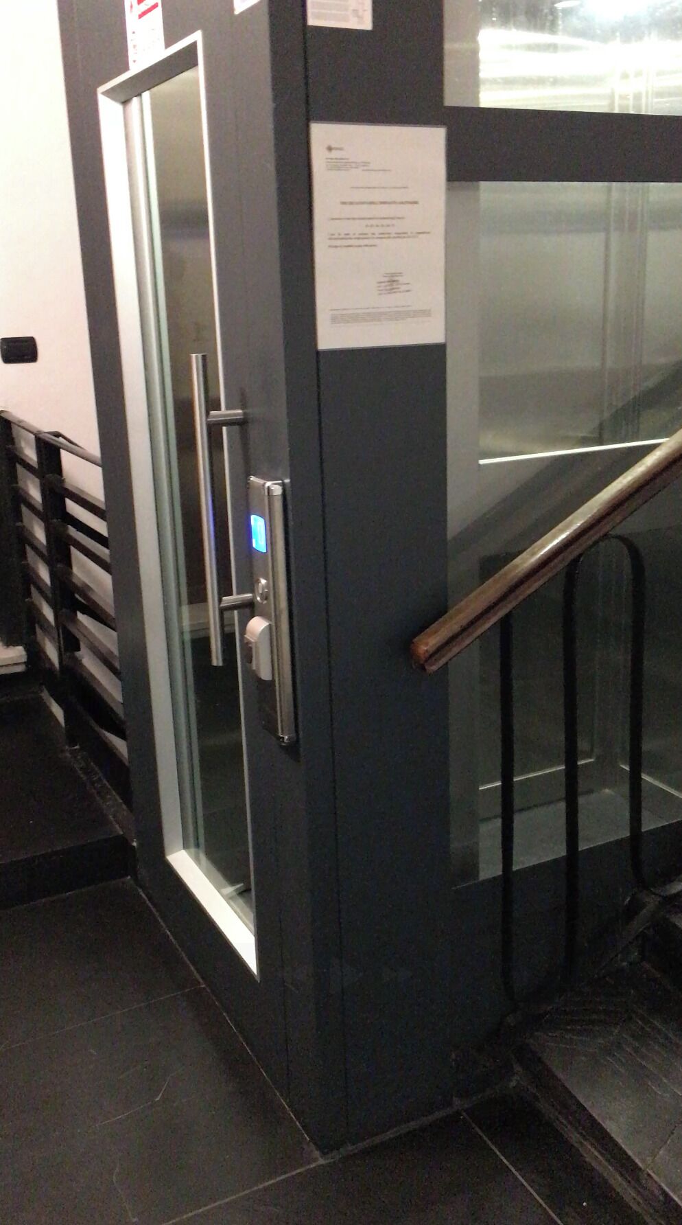 Un ascensore moderno con porta in vetro, display digitale e corrimano in metallo.