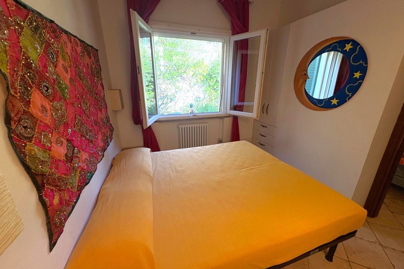 Ein Schlafzimmer mit einem Bett, einem Fenster, einer Heizung, einem runden Spiegel an der Wand und einem farbenfrohen Wandteppich.