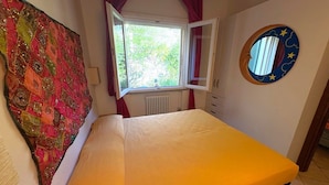 A bedroom with a bed, a window, a radiator, a wall-mounted round mirror, and a colorful tapestry. - Callistemo Apartment (Rio)