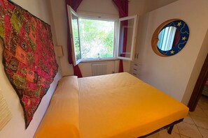 Ein Schlafzimmer mit einem Bett, einem Fenster, einer Heizung, einem runden Spiegel an der Wand und einem farbenfrohen Wandteppich.