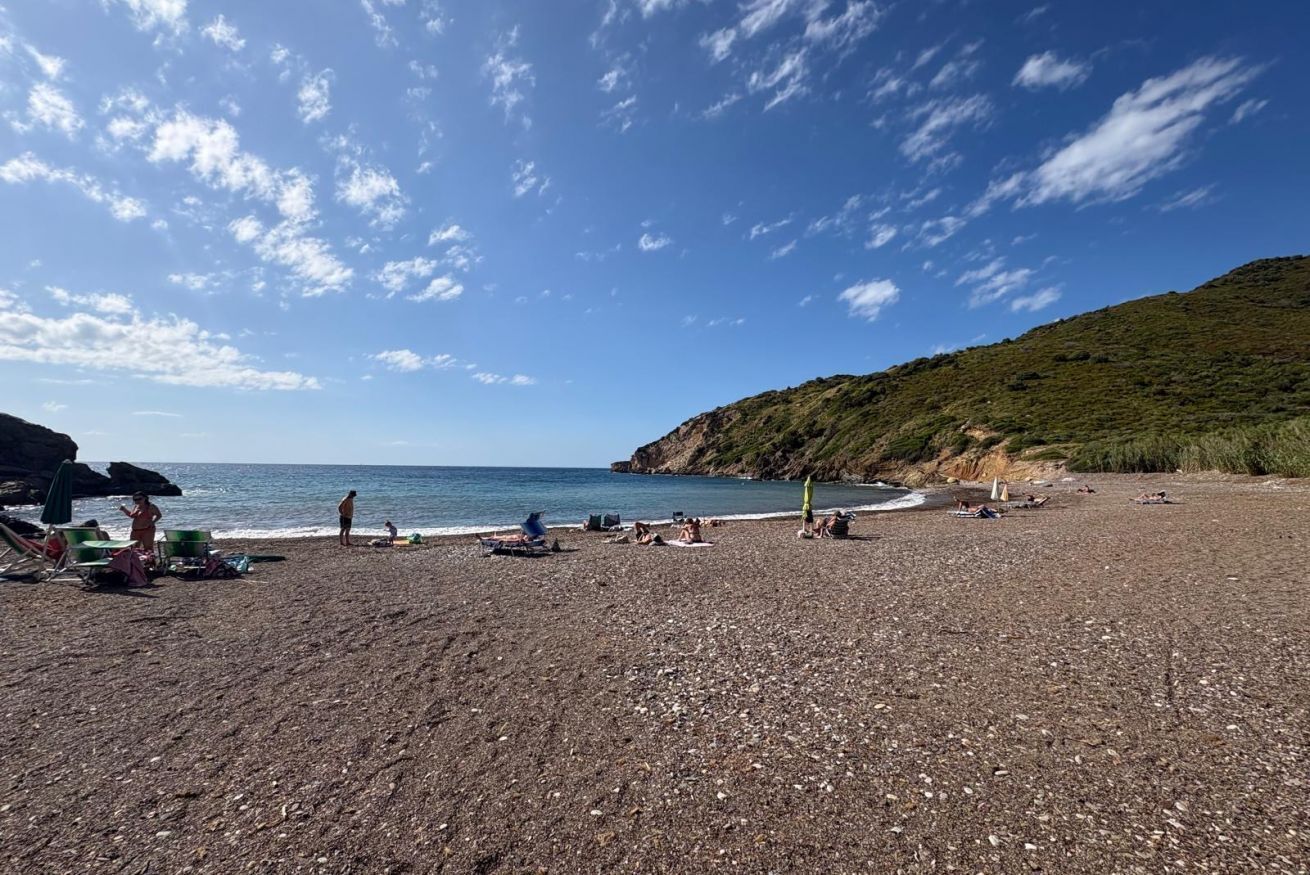 A pebbled beach with people sunbathing and swimming, a clear sky, and a hilly landscape in the background.