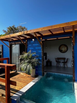 Zona de piscina con deck de madera, una planta en maceta y un área de descanso con mesa y sillas.