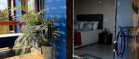 Un área de piscina moderna al aire libre con terraza de madera, una habitación con paredes de vidrio y una cama, y una planta en maceta.