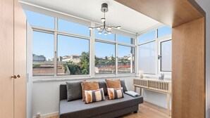 A modern living room with a grey sofa, multiple cushions, a wooden floor, and large windows offering a view of buildings and greenery. - GuestReady - Bright Studio in Bonfim District (Porto)