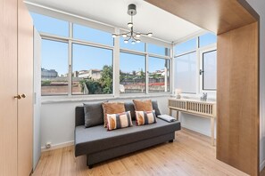 A modern living room with a grey sofa, multiple cushions, a wooden floor, and large windows offering a view of buildings and greenery. - GuestReady - Bright Studio in Bonfim District (Porto)