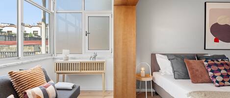A modern bedroom with a sofa, a bed, a bedside table, and a window with a view of buildings.